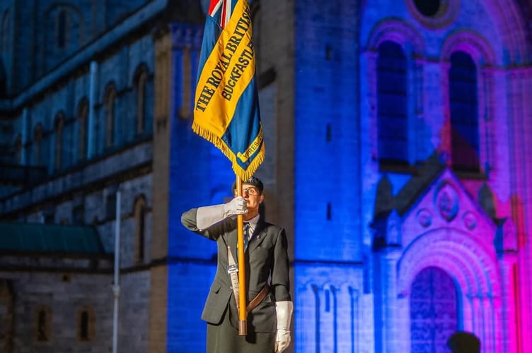 Buckfastleigh RBL standard bearer Julia Rouncefield in front of a lit up Buckfast Abbey to commemorate the 80th anniversary of VE Day. Photo courtesy of Buckfast Abbey