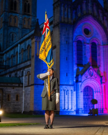 Buckfastleigh RBL standard bearer Julia Rouncefield in front of a lit up Buckfast Abbey to commemorate the 80th anniversary of VE Day. Photo courtesy of Buckfast Abbey